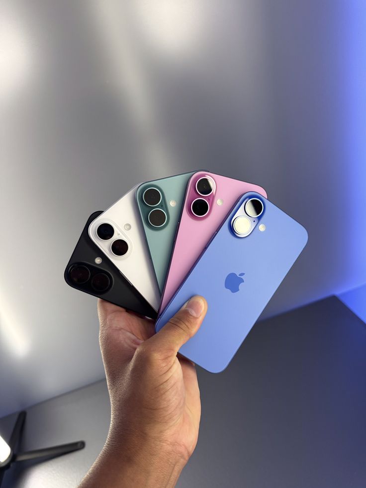 a hand holding four different colored iphones in front of a white wall with blue lighting