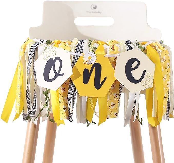 a yellow and black one banner with flowers on it is hanging from wooden legs in front of a white background