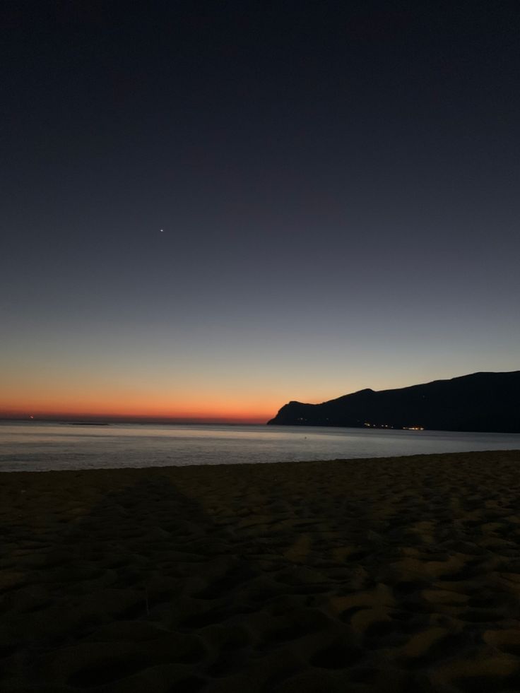 the sun is setting at the beach with mountains in the distance and water on the shore