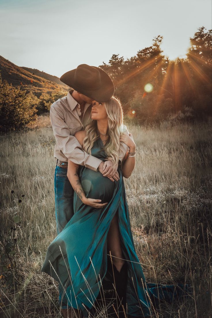 a pregnant woman in a blue dress and cowboy hat hugs her husband's belly