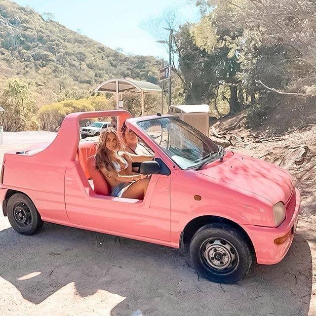 a dog sitting in the driver's seat of a pink convertible car with its front door open