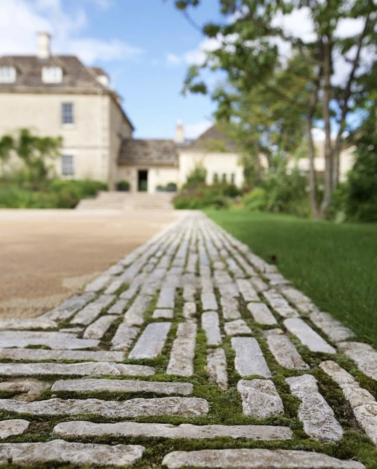 an old brick road with grass growing between it and a large house in the background