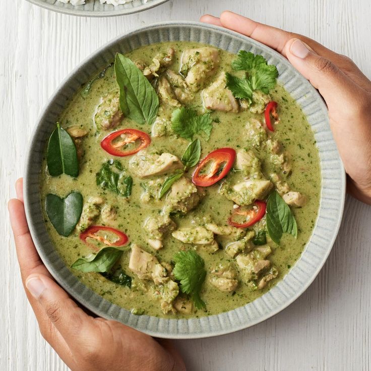 two hands holding a bowl of green curry with chicken and red bell peppers on the side