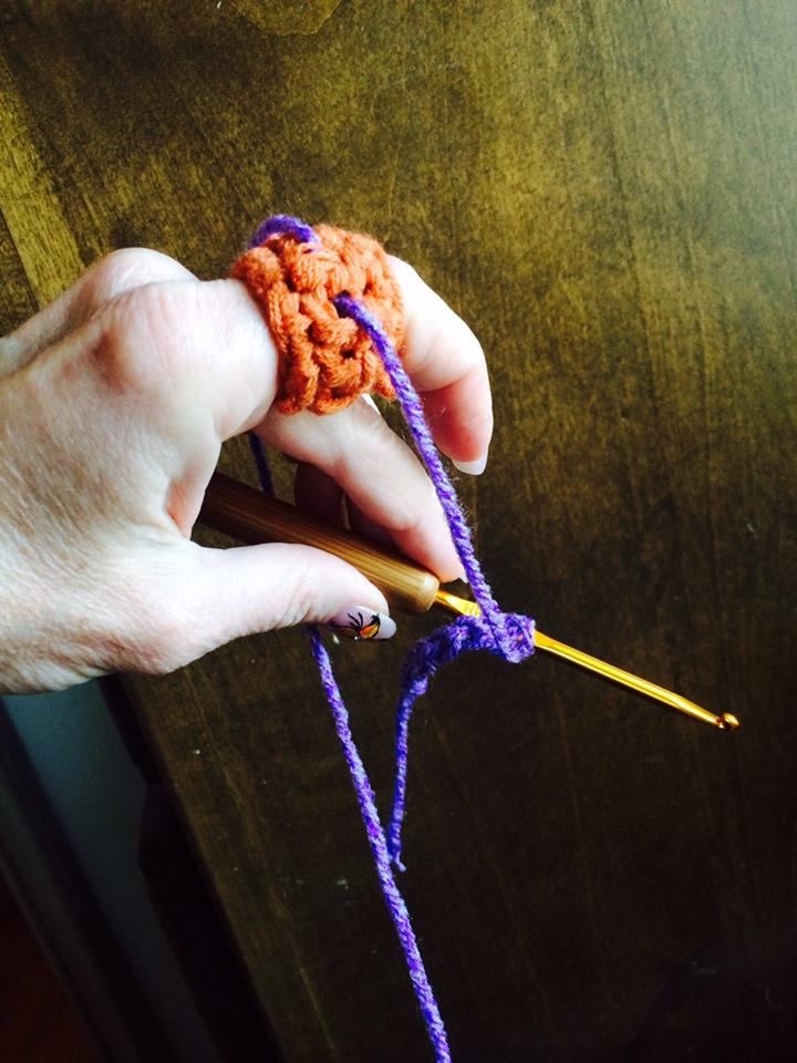 a hand holding a crochet hook with yarn on it and an orange ball of yarn in the other hand