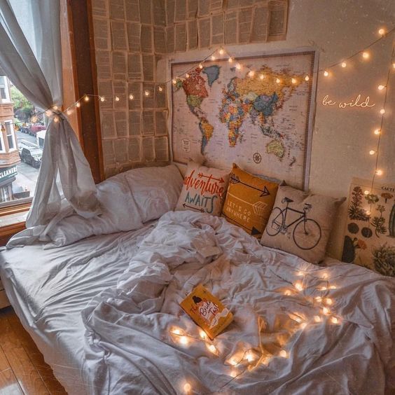 an unmade bed with white sheets and lights on the headboard is in front of a window