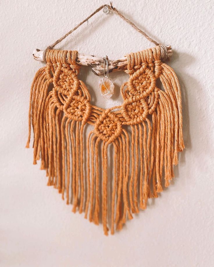 an orange macrame necklace hanging on a white wall next to a wooden stick