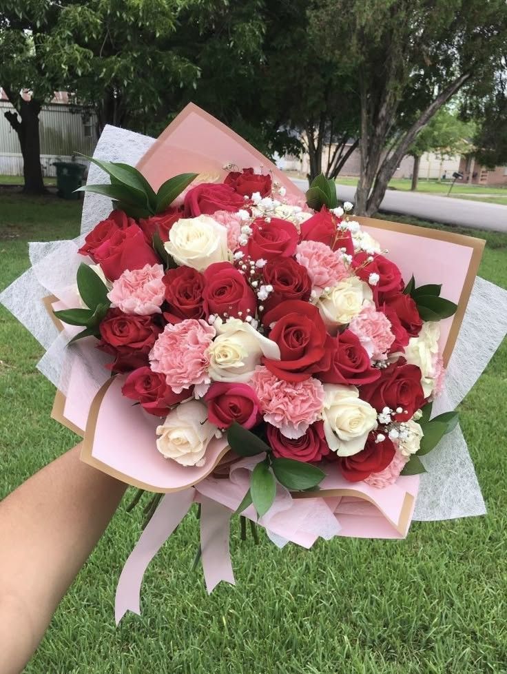 a bouquet of red and white flowers in someone's hand on the grass with trees in the background