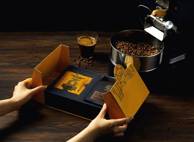 two hands opening an open box with coffee beans in it on a wooden table next to a metal pot