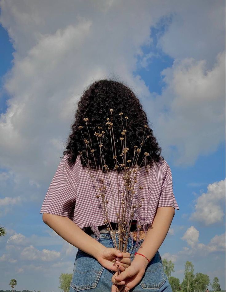 a woman with long curly hair is holding flowers in her hands and looking at the sky