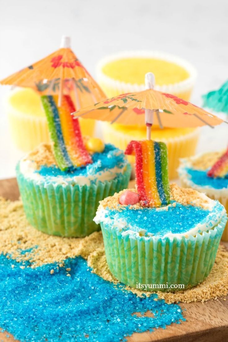 cupcakes decorated with rainbow colored frosting and an umbrella are on a wooden board