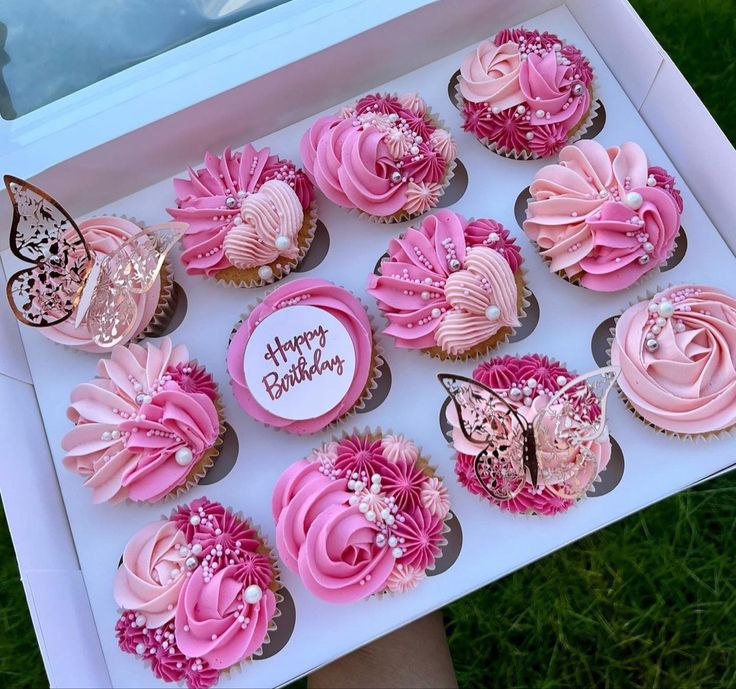 a box filled with pink cupcakes on top of a grass covered field next to a person's hand