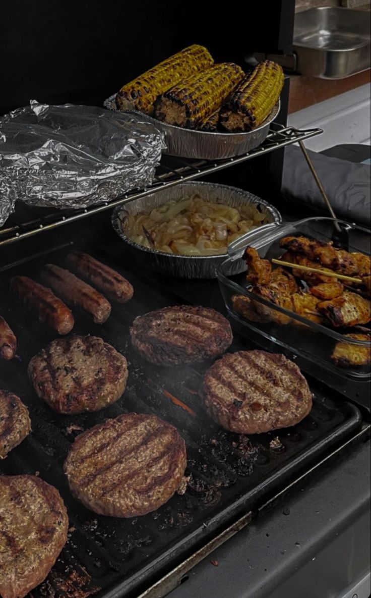 hamburgers, corn on the cob and other foods cooking on an outdoor grill