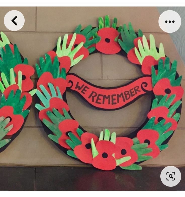 handprinted wreath with red flowers and green leaves on brick wall next to sign that says, we are remembrance