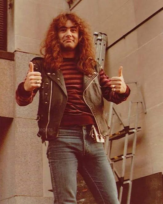 a man with long hair giving the thumbs up while standing in front of a building