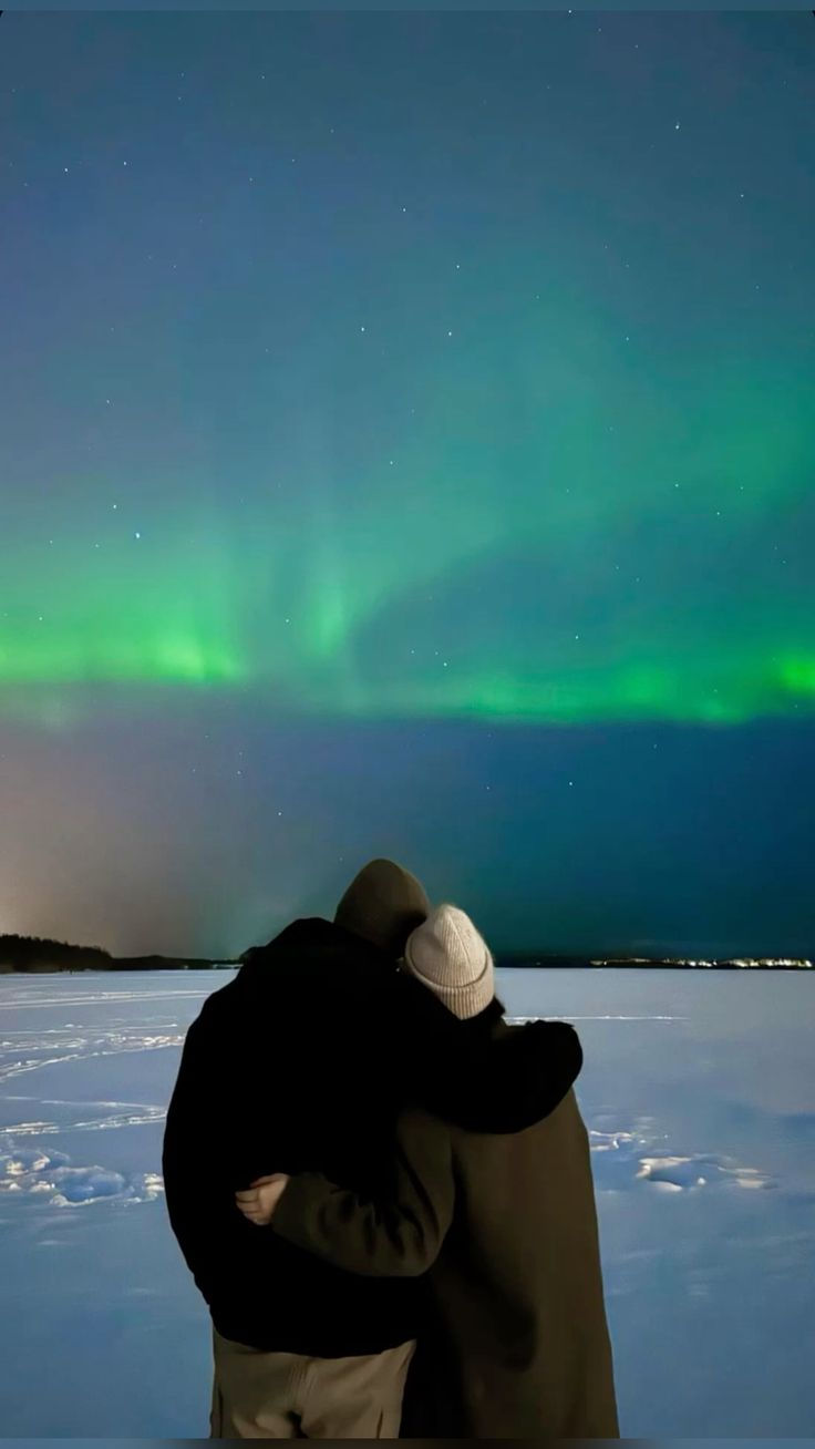 two people standing in the snow looking at an aurora bore above them, with their arms around each other