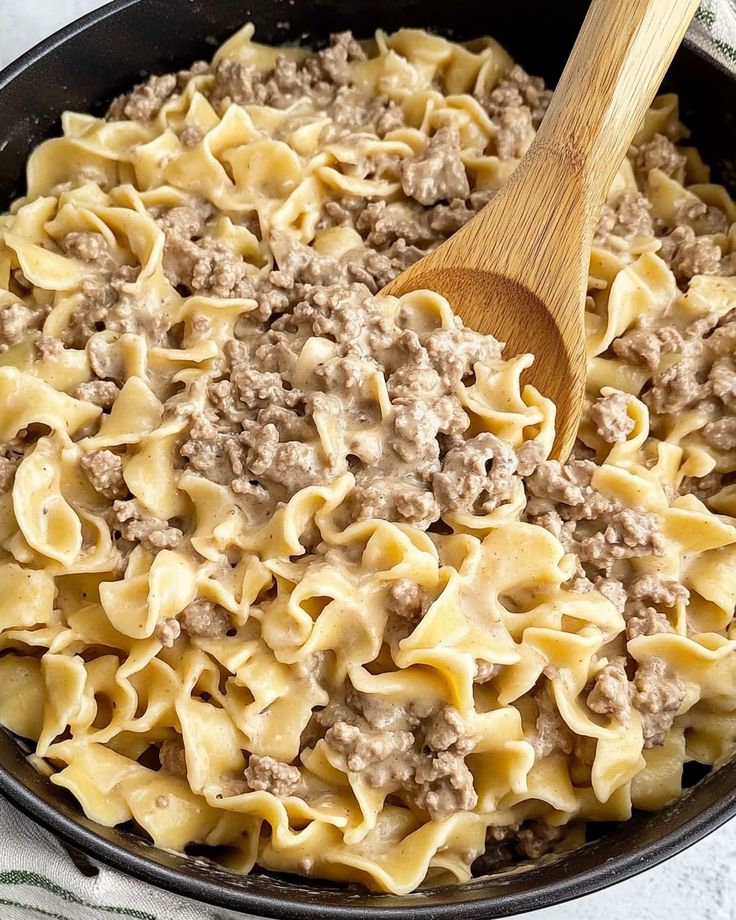 a skillet filled with pasta and meat on top of a white cloth next to a wooden spoon