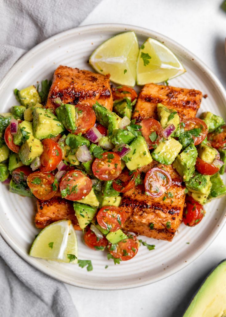 a white plate topped with chicken, avocado and tomato salad next to sliced limes