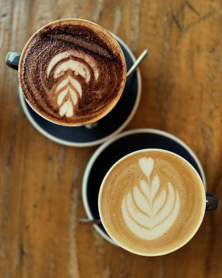 two cups of cappuccino with designs on them sitting on a wooden table