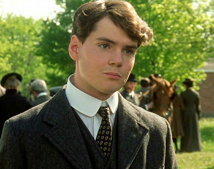 a man wearing a suit and tie standing in front of some people with horses behind him