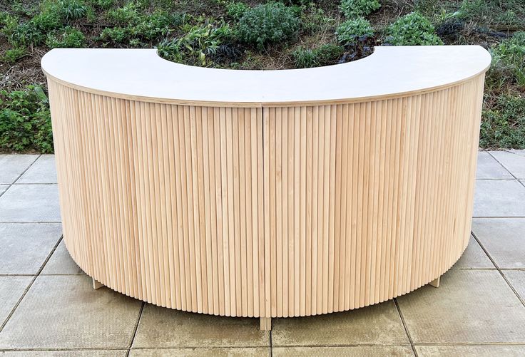 a curved wooden table sitting on top of a tile floor next to a green bush