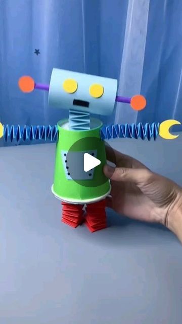 a hand holding a green and red robot toy on top of a gray table next to a blue curtain