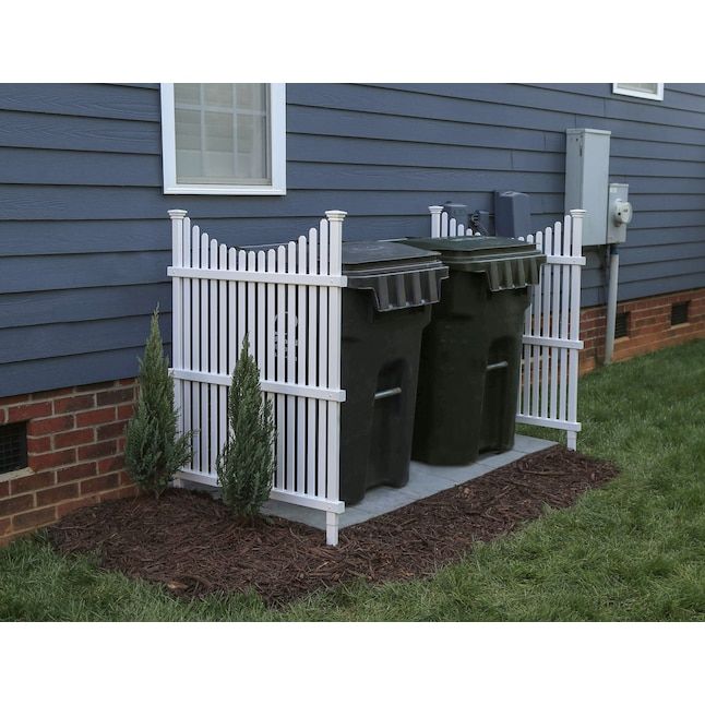 two trash cans sitting in front of a white picket fence on the side of a house