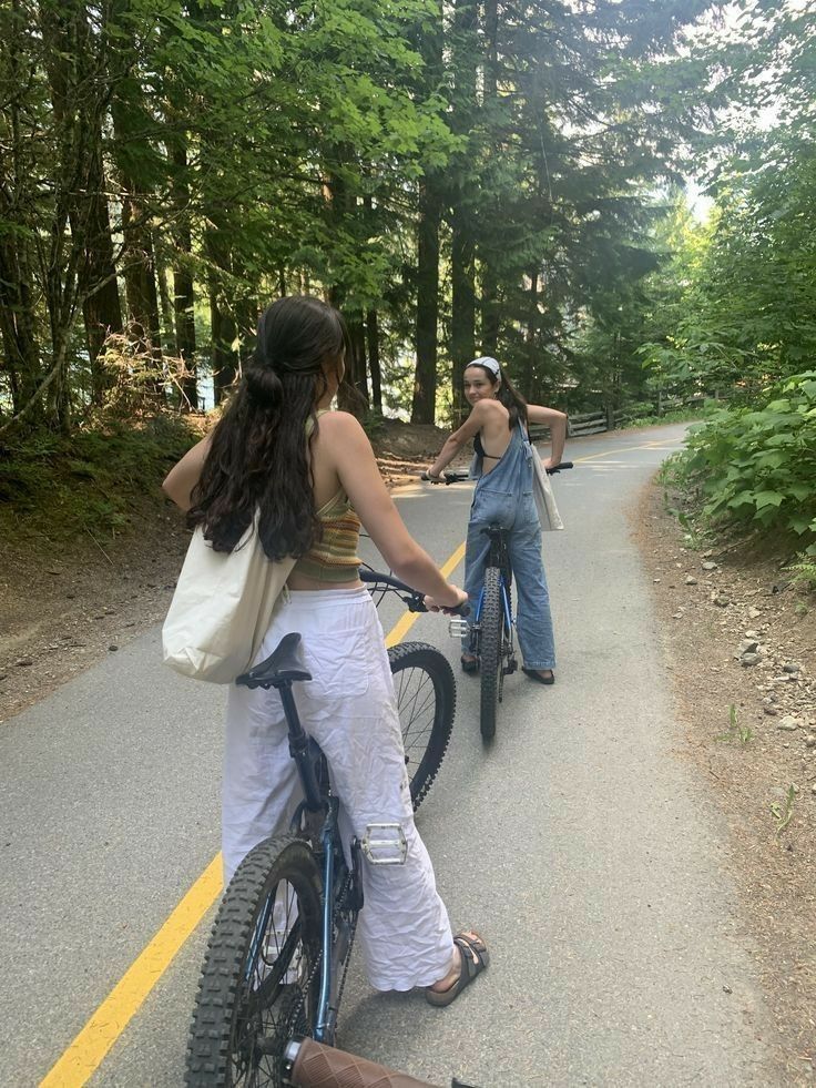 two women riding bikes on the side of a road with trees in the back ground