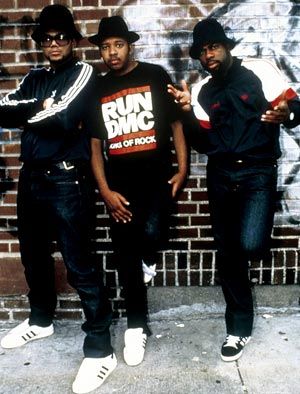 three young men are standing in front of a brick wall with graffiti on it and one is wearing a run d m c t - shirt