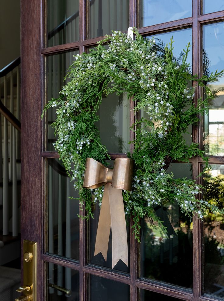 a christmas wreath hanging on the front door of a house with gold bow and greenery