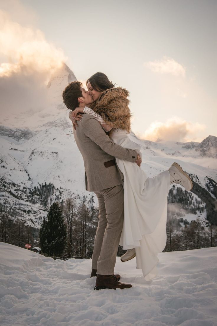 a bride and groom kissing in the snow with mountains in the background