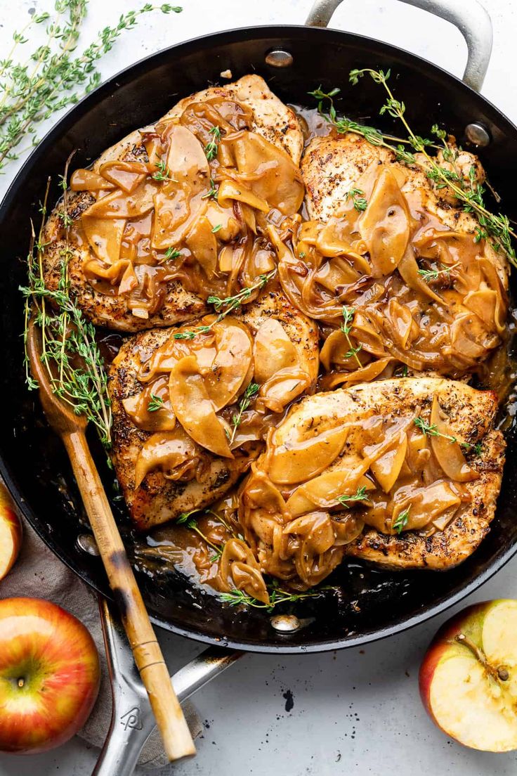 chicken with apple sauce in a skillet next to sliced apples and herbs on the side