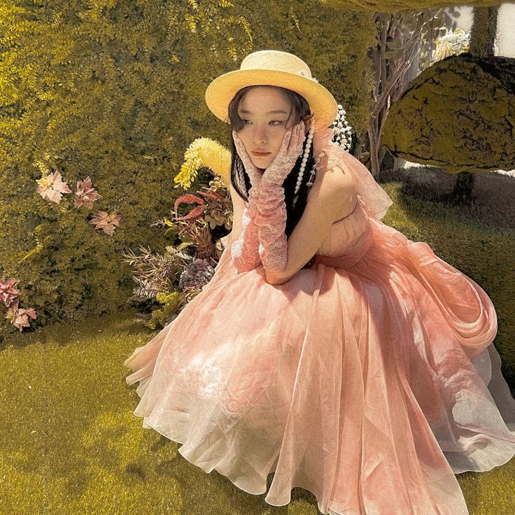 a woman in a pink dress and hat sitting on the grass with her hands up to her face