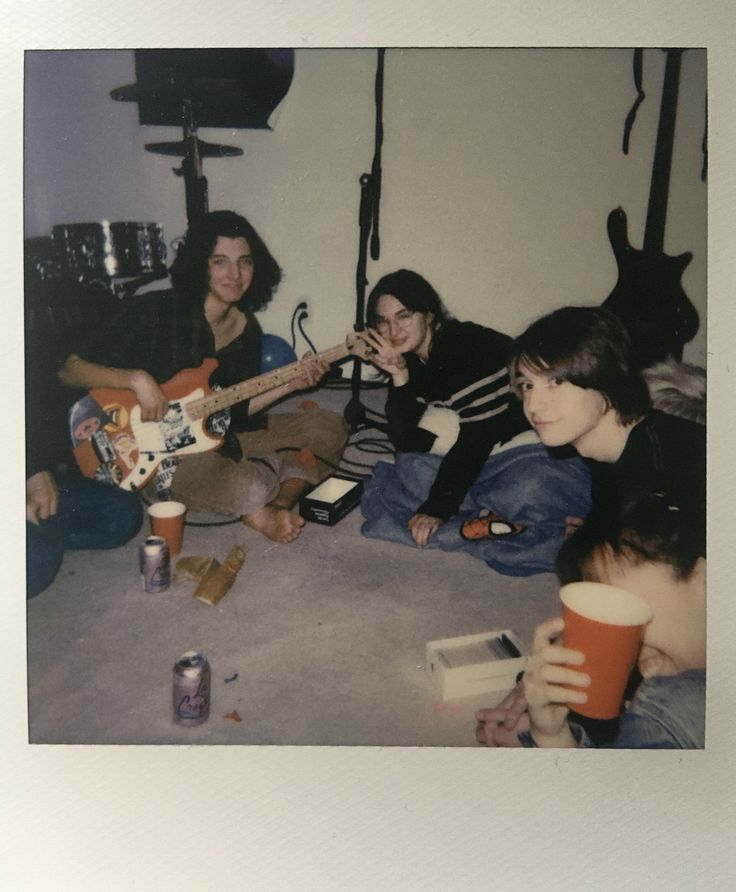 four people sitting on the floor playing guitars and drinking beer while one person holds a guitar