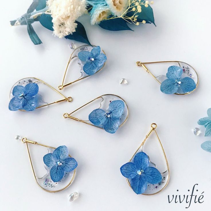 blue flowers are hanging from gold earwires on a white table next to dried flowers