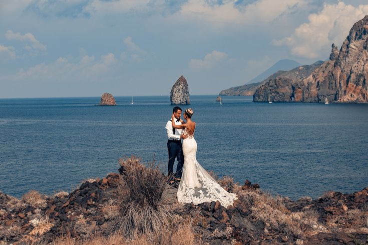 a bride and groom standing on the rocks by the ocean with their arms around each other