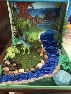 an open cardboard box with a dinosaur in the grass and rocks on the ground next to it