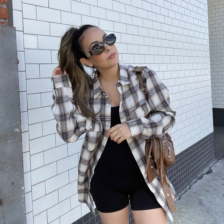 a woman in black shorts and plaid shirt standing next to a white brick wall with her hand on her head