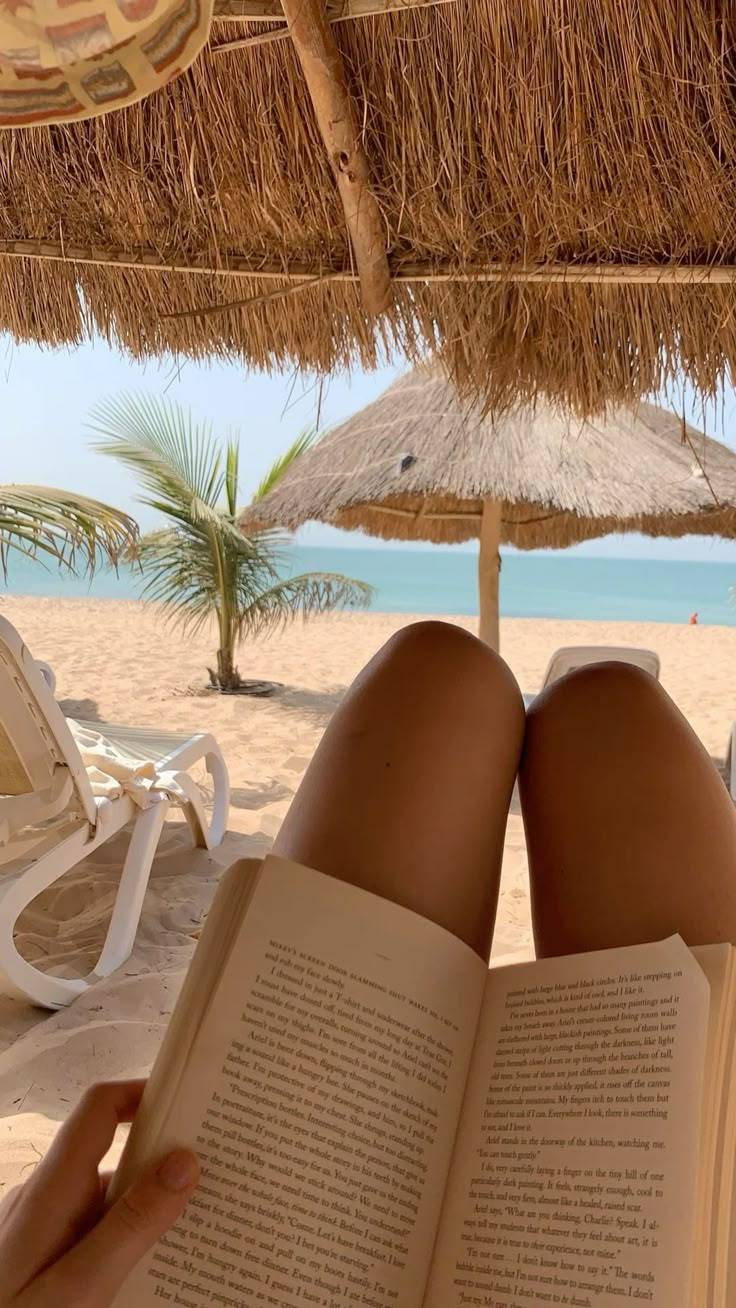 a person reading a book on the beach under an umbrella with their feet propped up