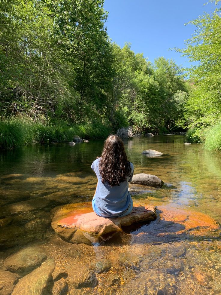 a woman sitting on top of a rock in the middle of a river surrounded by trees