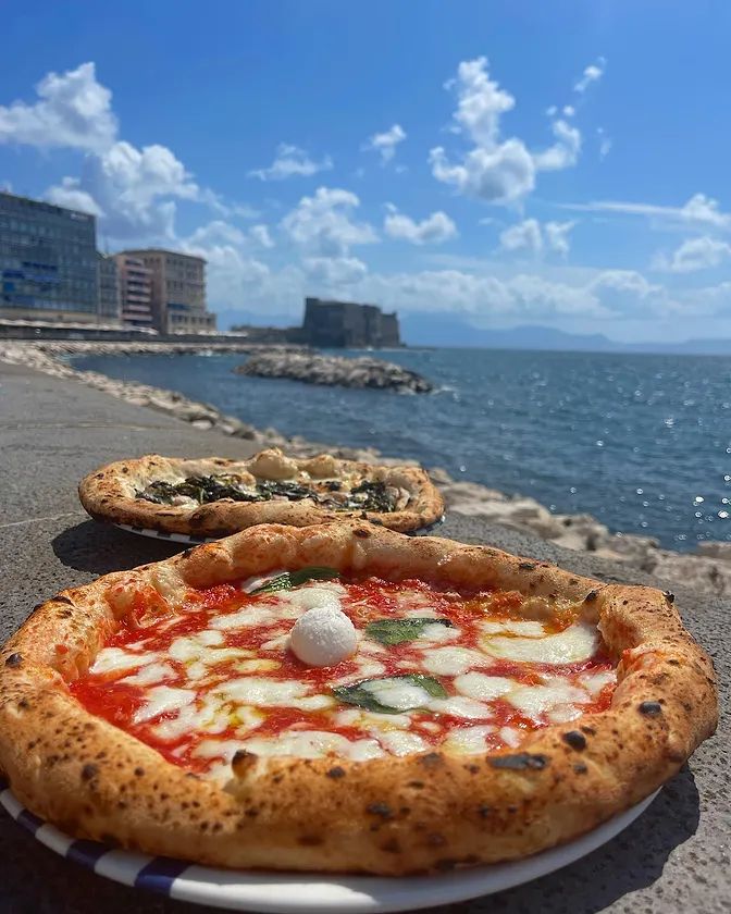 two pizzas sitting on top of a white plate near the ocean with buildings in the background