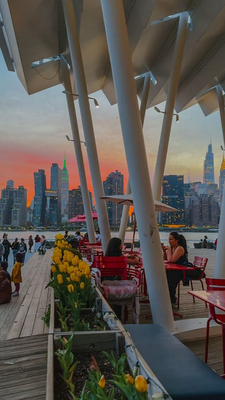 people are sitting at tables on the pier near the water and skyscrapers in the background