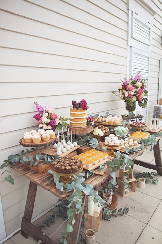 a table topped with cakes and desserts on top of a wooden table covered in greenery