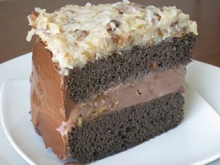 a piece of chocolate cake with frosting and nuts on top sitting on a white plate