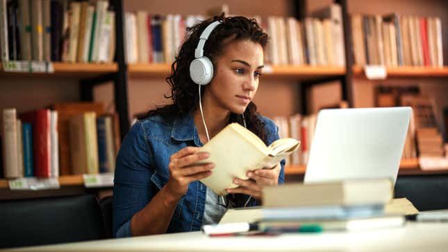 a woman sitting in front of a laptop computer wearing headphones and reading a book