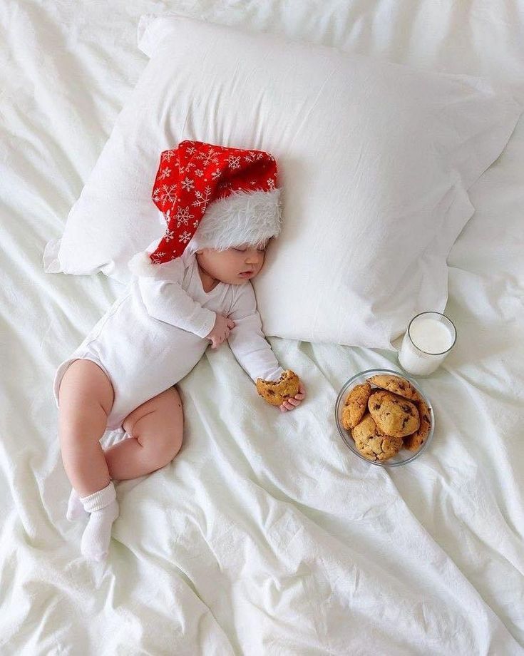 a baby laying on a bed with cookies and milk in front of it while wearing a santa hat