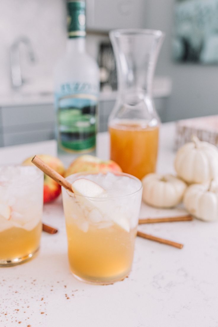 two glasses filled with drinks sitting on top of a counter next to apples and cinnamon sticks