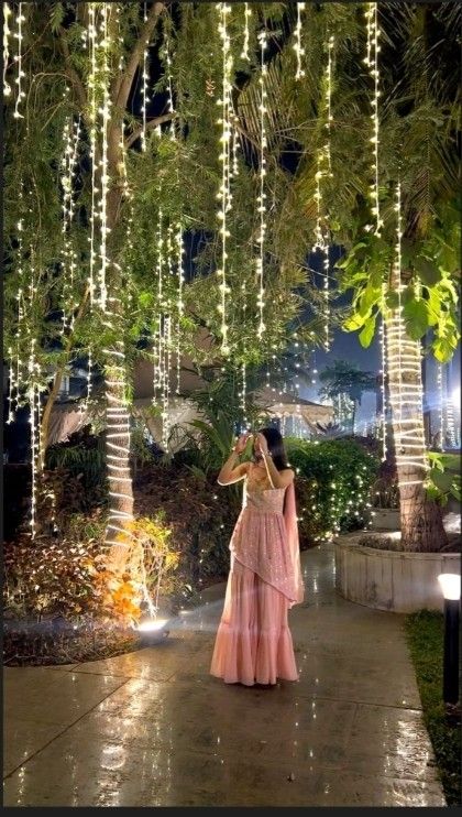 a woman in a pink dress is standing under some trees with lights hanging from them