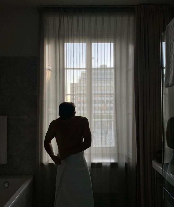 a man standing in front of a window with his back turned to the camera while wearing a towel