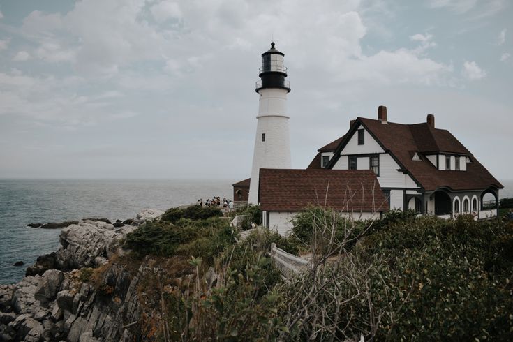 a white house with a black roof and a light house on top of it near the ocean