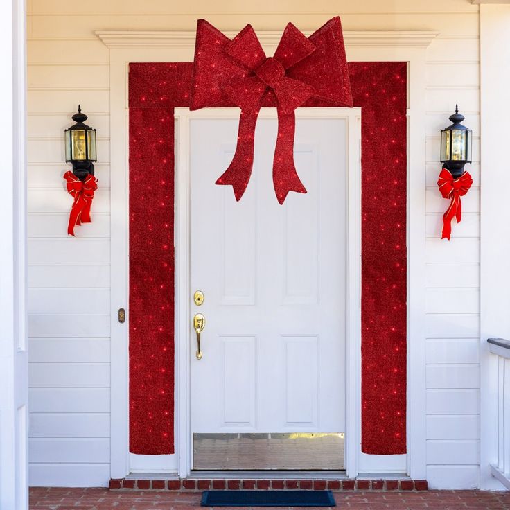 a red bow on the front door of a white house with two lanterns and lights